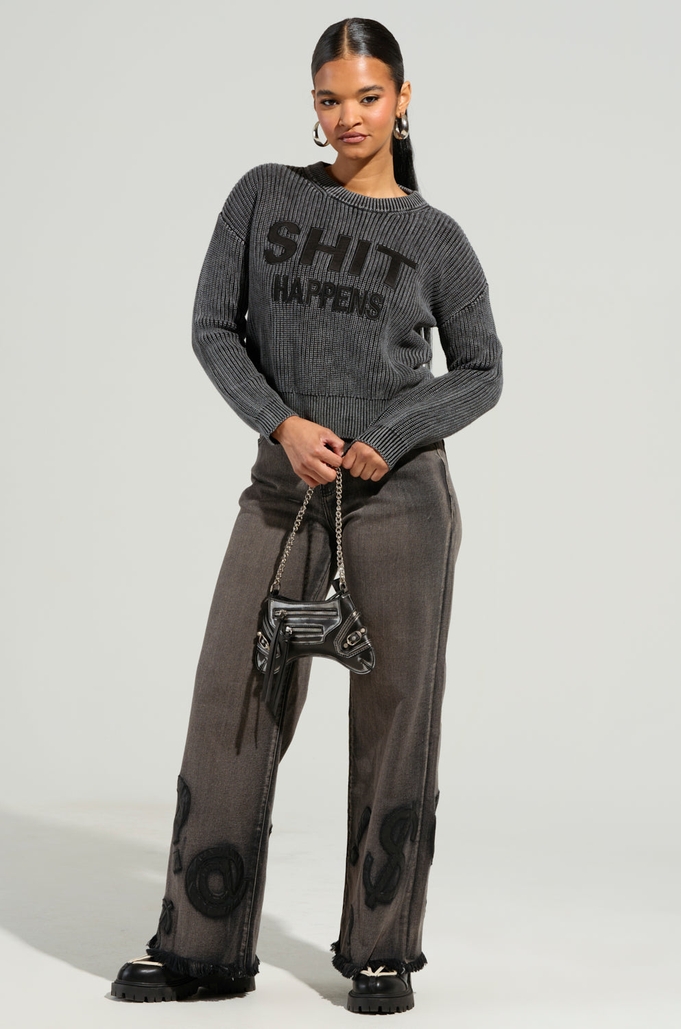 A person stands confidently wearing the READ MY LIPS PATCHWORK DENIM JEANS, accessorized with a gray sweater featuring bold black text, hoop earrings, a small black handbag, and faux leather patches. They pose against a plain background.