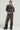 A person stands confidently wearing the READ MY LIPS PATCHWORK DENIM JEANS, accessorized with a gray sweater featuring bold black text, hoop earrings, a small black handbag, and faux leather patches. They pose against a plain background.