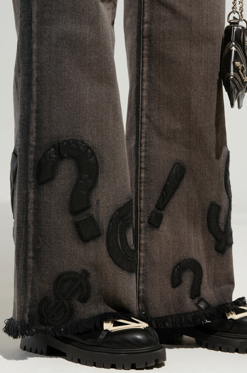 A detailed view of someone in dark denim featuring the "READ MY LIPS PATCHWORK DENIM JEANS," highlighted by embroidered faux leather patches with large black question and exclamation marks. The frayed hems complement black loafers adorned with fringe details, while a small black handbag is subtly visible.