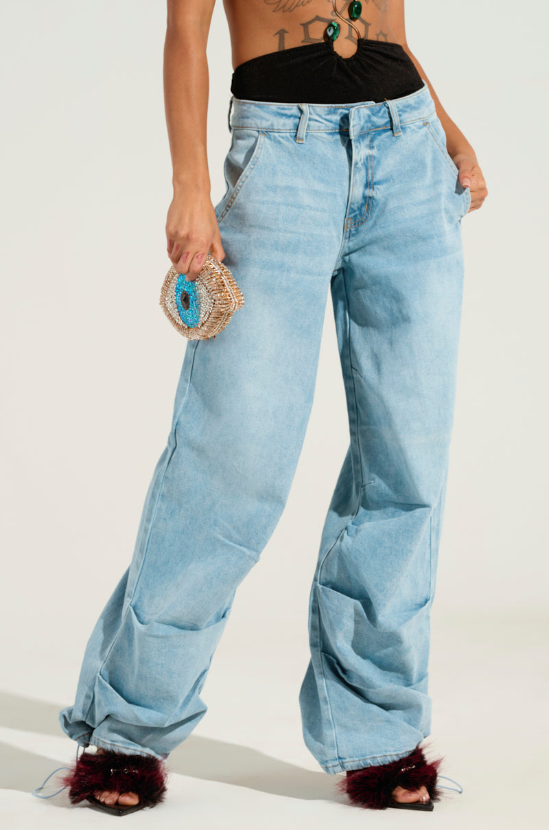 A person is wearing LUCKY LUCKY WIDE LEG DENIM PANT in light blue with a wide leg silhouette, paired with a black crop top and carrying a small woven handbag featuring a blue pattern. On their feet are maroon furry slippers, all set against a plain white background.