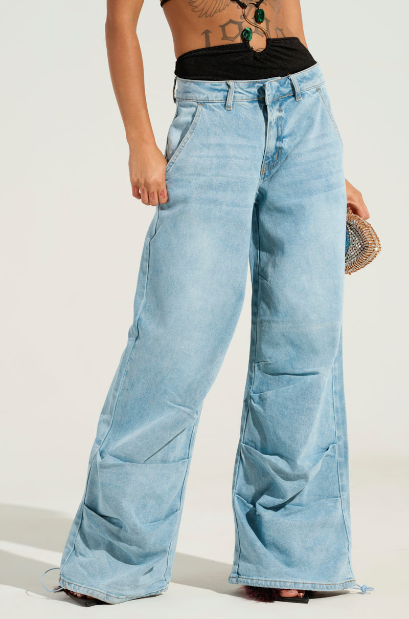 A person wearing Lucky Lucky's LUCKY LUCKY WIDE LEG DENIM PANT in light blue, characterized by a high-rise waist and relaxed fit, holds a small woven bag. They sport a black top wrapped around the chest, revealing an abdominal tattoo against a minimalist and light-colored backdrop.