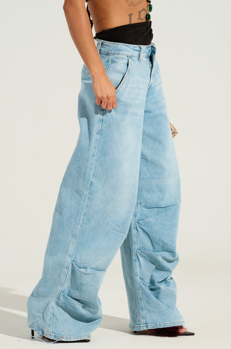 Wearing the LUCKY LUCKY WIDE LEG DENIM PANT in an oversized light blue style with a wide leg silhouette, a person pairs them with a black top and poses with one hand in their pocket. The high-rise jeans extend to the floor, partially concealing their high-heeled shoes.