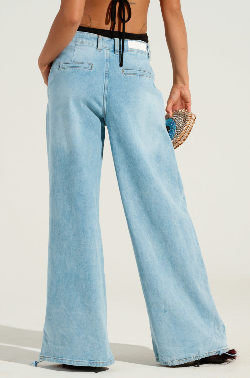 A person wearing light blue, wide-legged jeans with a high-rise waist and a black halter top holds a woven round clutch. The photo is taken from behind, highlighting the LUCKY LUCKY WIDE LEG DENIM PANT's fit and style against a plain light background.