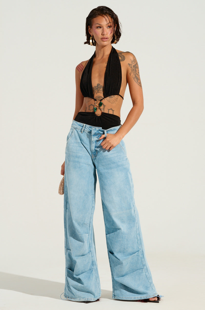 A person with short hair is dressed in a black halter top and LUCKY LUCKY WIDE LEG DENIM PANT. Displaying tattoos on their arm, they pose against a neutral backdrop, holding a small woven bag.