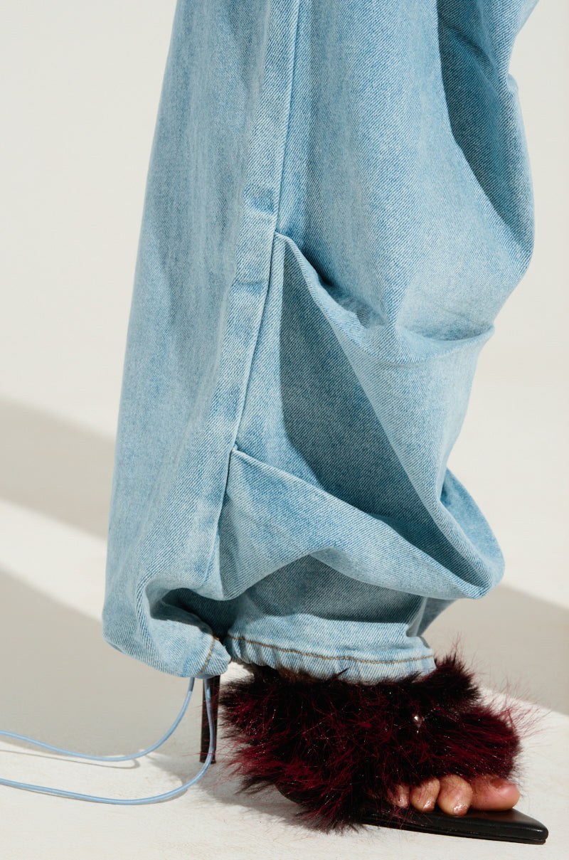 A close-up of a person wearing the LUCKY LUCKY WIDE LEG DENIM PANT in a loose, light blue design with a high-rise waist, highlighting only the wide-leg silhouette. The pants are matched with open-toed sandals adorned with fluffy, dark red fur against a plain light background.