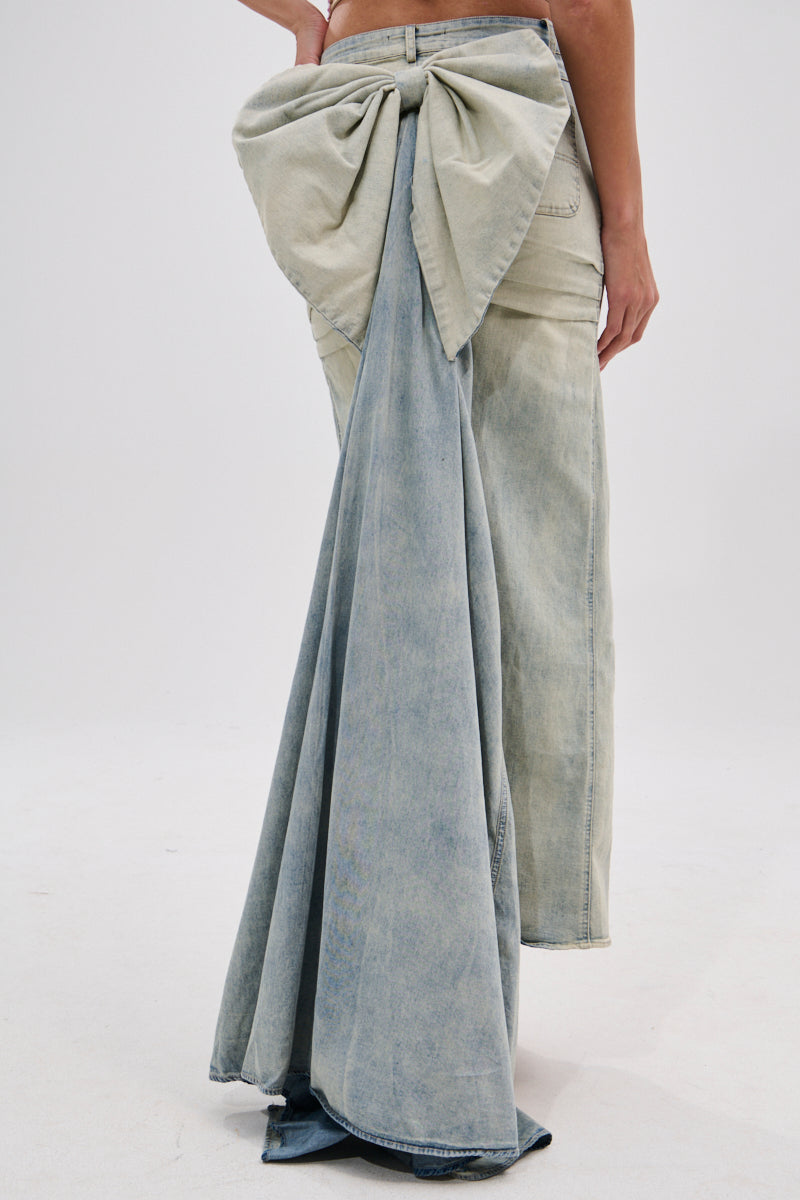 Someone stands against a plain light background wearing the OH THE DRAMA BACK BOW DENIM MAXI SKIRT WITH TRAIN, featuring light-wash denim, a dramatic back bow, and a floor-grazing train.