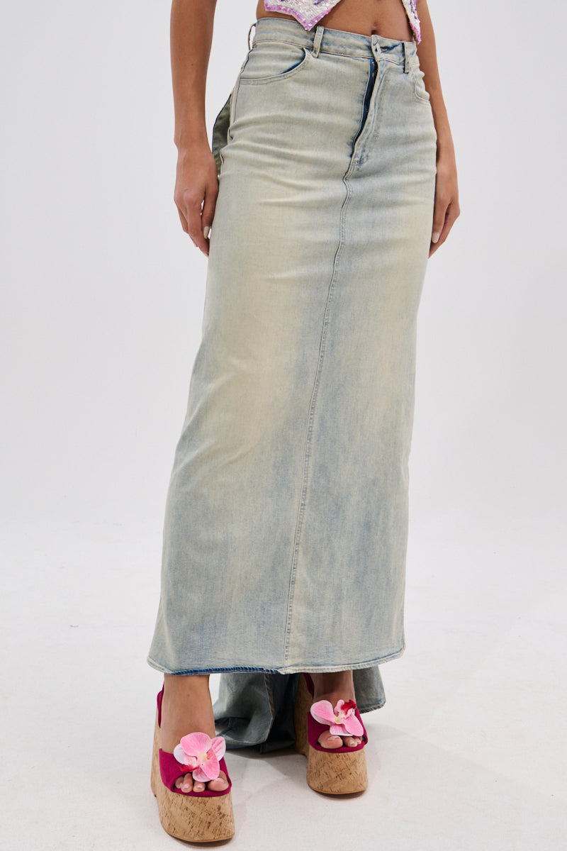 The lower half of a person is shown wearing the OH THE DRAMA BACK BOW DENIM MAXI SKIRT WITH TRAIN in faded light blue, styled with pink flower-adorned platform sandals, standing against a plain white background.