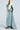 Wearing the OH THE DRAMA BACK BOW DENIM MAXI SKIRT WITH TRAIN, a person pairs this high-rise, light blue denim skirt featuring back pleats with a cropped knit sweater and zebra-patterned high-heeled boots, against a plain, light background.