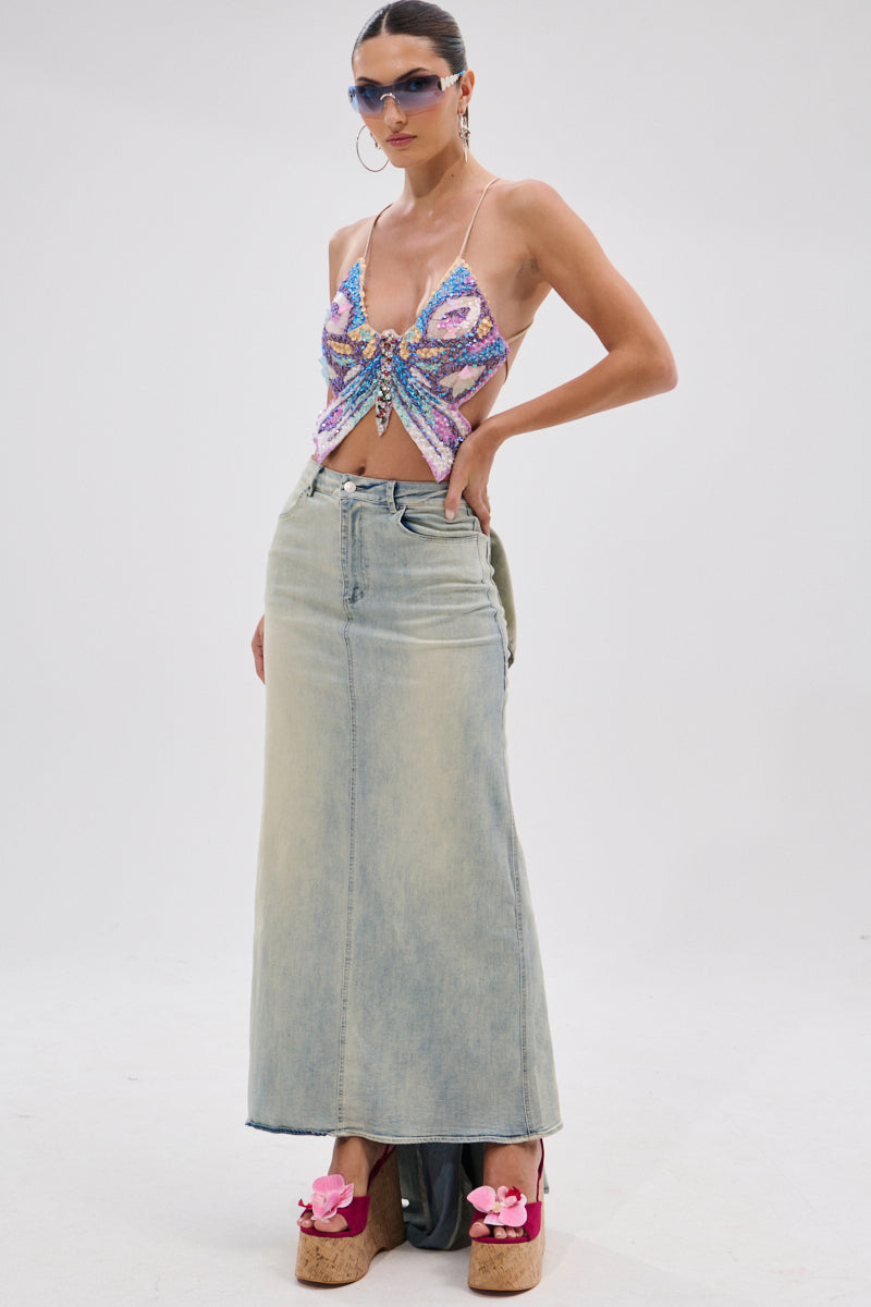 A woman wearing sunglasses, a butterfly halter top, platform sandals with pink flowers, and the OH THE DRAMA BACK BOW DENIM MAXI SKIRT WITH TRAIN stands against a plain white background.