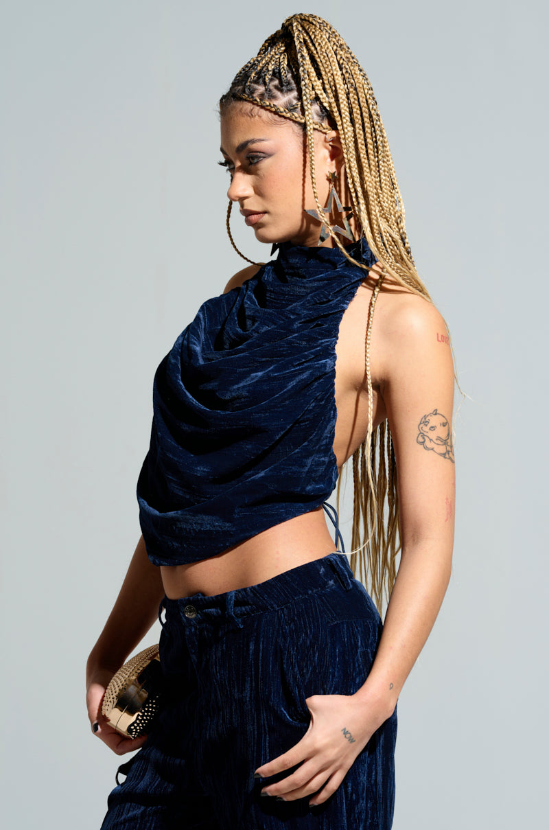 A person with long braided hair is wearing the FEELING TEMPTED HALTER NECK TIE BACK TOP in a dark blue color, along with matching pants. They hold a small clutch purse, showcasing tattoos on their arm. The outfit features an open-back design and is set against a plain, light-colored background. 1