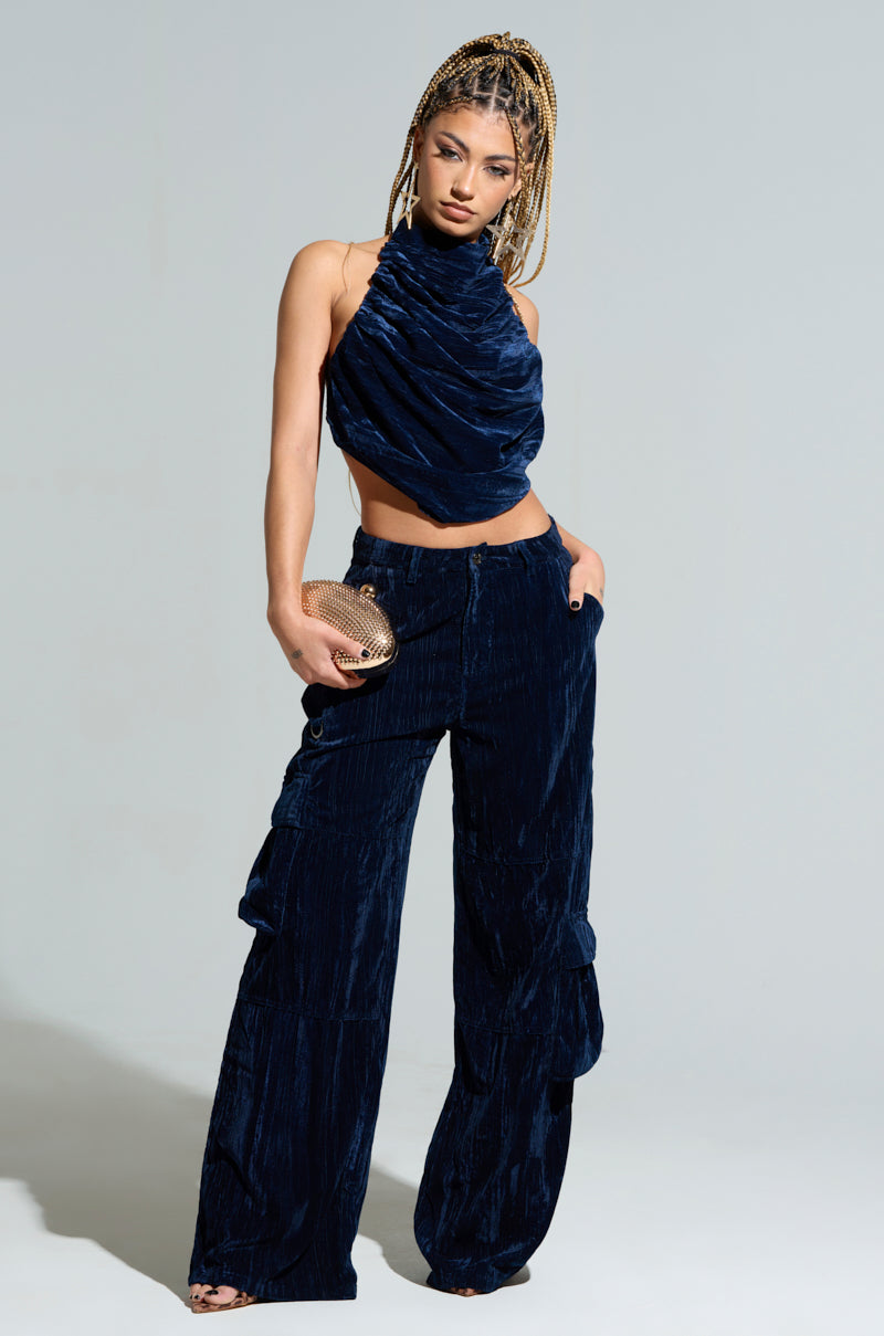 A woman poses confidently in the FEELING TEMPTED HALTER NECK TIE BACK TOP made of blue crushed velvet, featuring an open-back design. She complements it with matching wide-leg pants and holds a small gold clutch in one hand, her long braided hair cascading gracefully against the neutral gray backdrop. 3