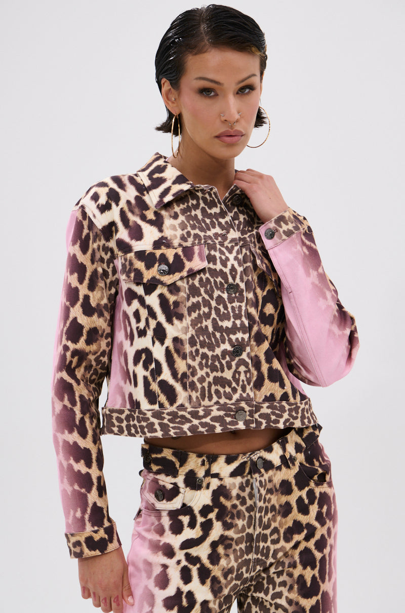 A woman poses confidently in the COCO LEOPARD PRINT DENIM JACKET and matching pants, touching her collar. She wears slicked-back hair, hoop earrings, and displays a bold expression against a plain background.