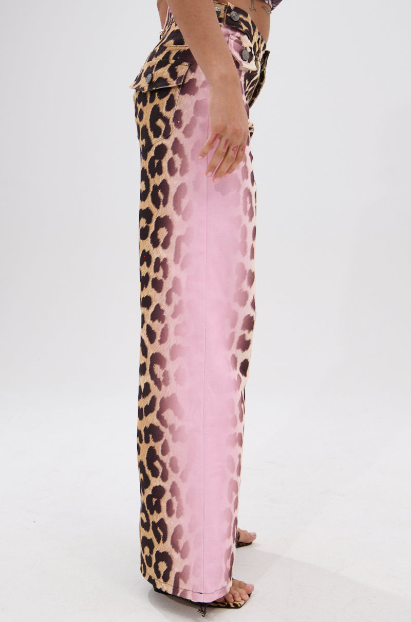 A person stands sideways wearing COCO LEOPARD PRINT DENIM high-waisted pants, featuring a bold leopard print that blends into pink on the inner leg, paired with open-toe heels against a plain white background.