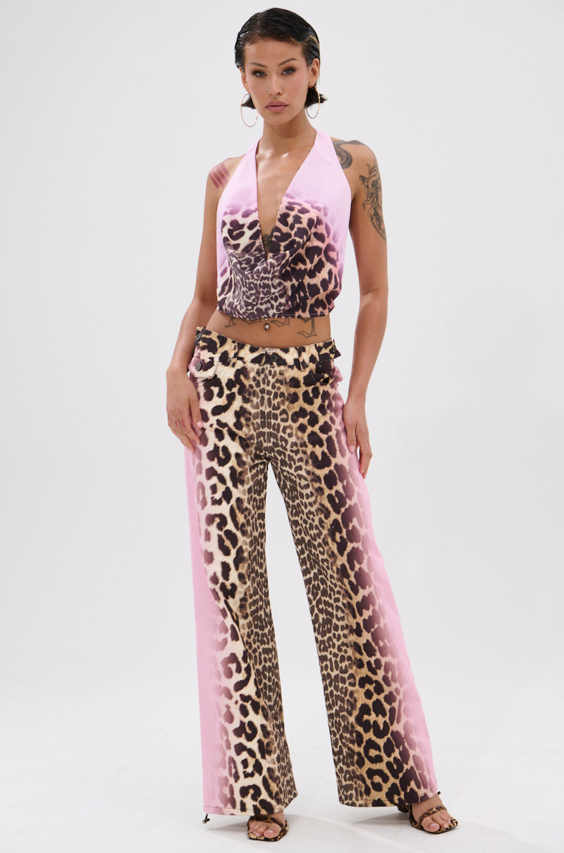 A person stands confidently in the COCO LEOPARD PRINT DENIM, featuring a sleeveless halter top and high-waisted flared pants with pink and leopard print, accessorized with hoop earrings and sandals against a white background.