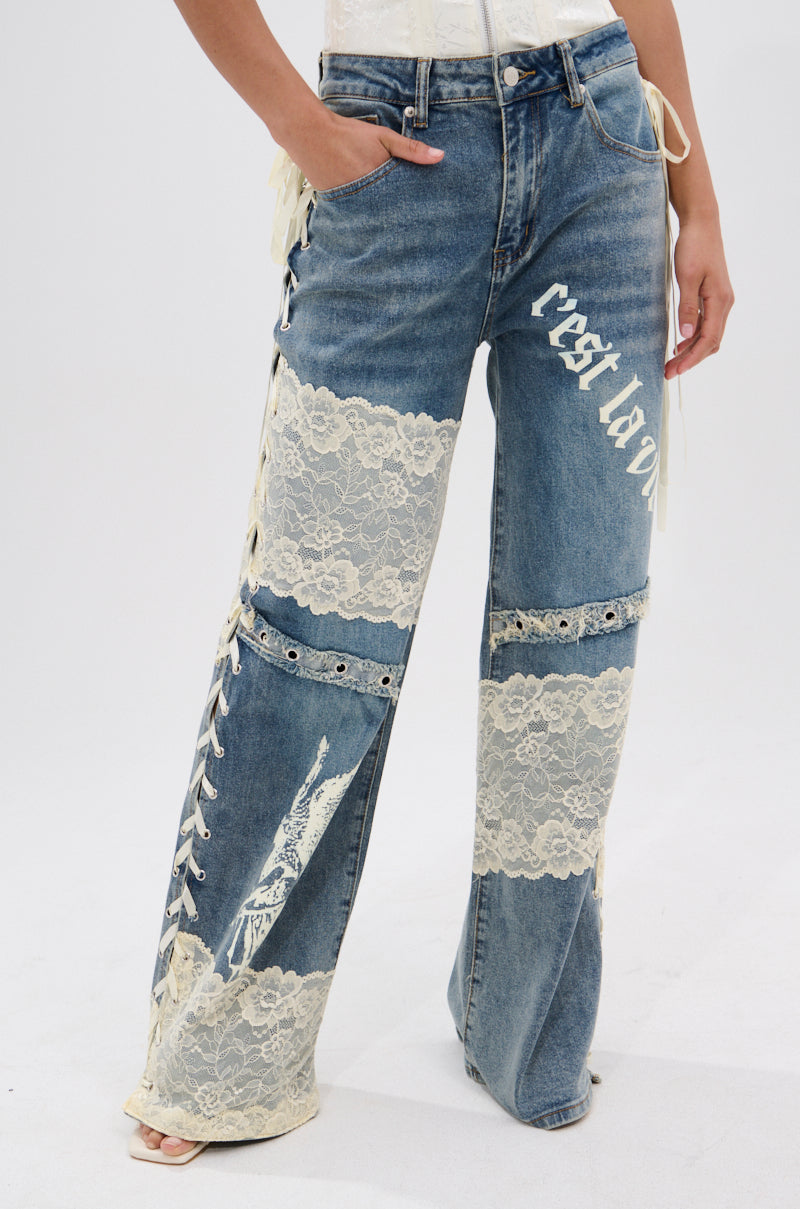 Wearing FEMME FATALE PRINTED AND LACE UP DETAIL DENIM JEANS, featuring cream floral lace panels, side lace-up details, and a white "C'est La Vie" print on one leg; only the lower body is shown against a light background.