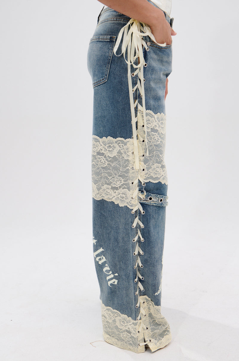 A person wears FEMME FATALE PRINTED AND LACE UP DETAIL DENIM JEANS—blue denim with cream floral lace panels, side lace-up with cream ribbons and eyelets—while standing against a plain white background.