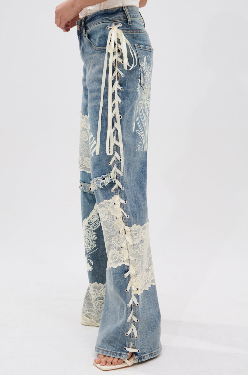 A person wears FEMME FATALE PRINTED AND LACE UP DETAIL DENIM JEANS with white floral lace paneling and cream lace-up sides, paired with open-toed shoes. The wide-leg jeans create a chic, romantic look.