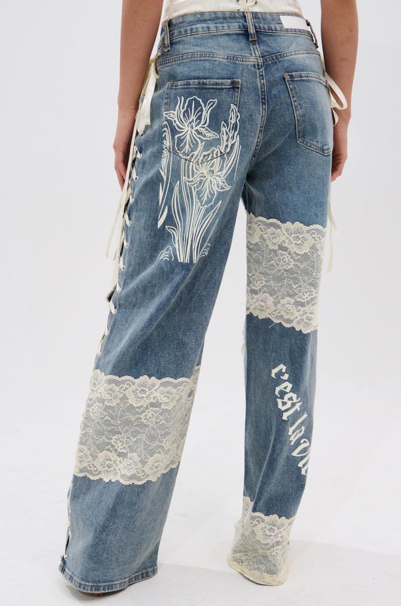 Someone wears FEMME FATALE PRINTED AND LACE UP DETAIL DENIM JEANS with white floral lace panels, side lace-up details, and a "crest a love" graphic. The photo, taken from behind, highlights the bold design.