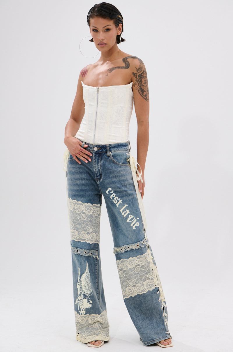 A woman poses against a white background in a strapless white corset top and Femme Fatale Printed and Lace Up Detail Denim Jeans with cream floral lace panels and “c’est la vie.” She sports tattoos, hoop earrings, and braided hair.