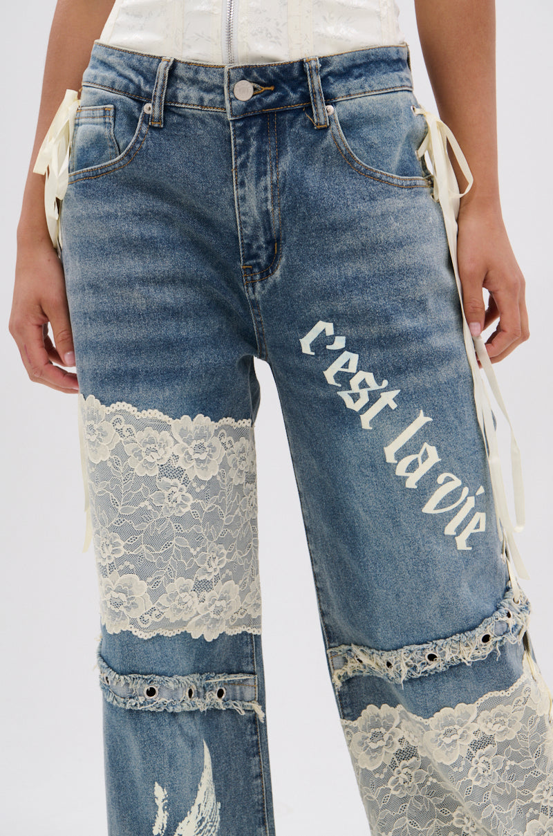 Someone wearing FEMME FATALE PRINTED AND LACE UP DETAIL DENIM JEANS, featuring blue denim with white floral lace panels, "c'est la vie" print on one thigh, and decorative side ribbons for added charm.