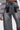 A person wears the JACQUELINE CUTOUT DENIM PANTS—high-rise, vintage-washed gray denim with large black rectangular patches above the back pockets—and a grey top with sheer panel sleeves. The image highlights the back of the jeans.
