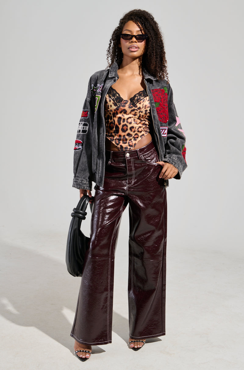 A woman wearing sunglasses poses in a leopard print top, a grey jacket with patches, and shiny high-rise faux leather trousers in burgundy. She holds a black handbag and stands against a plain background. 1