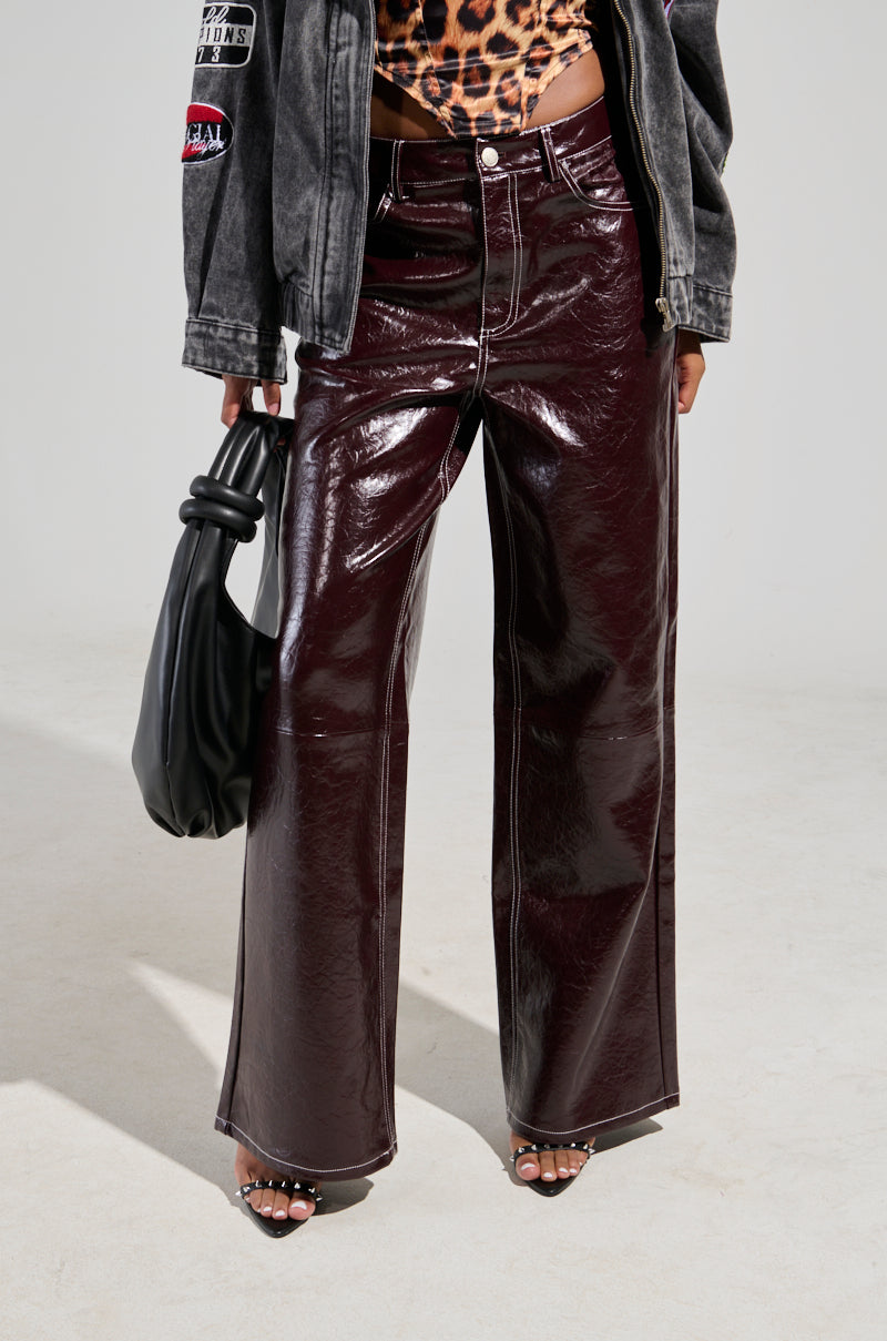 A person is dressed in the "START WITH ME FAUX LEATHER TROUSER IN BURGUNDY," paired with a leopard print top and a gray jacket. They are holding a black handbag and wearing open-toed shoes, standing against a plain, light-colored floor and wall. 0
