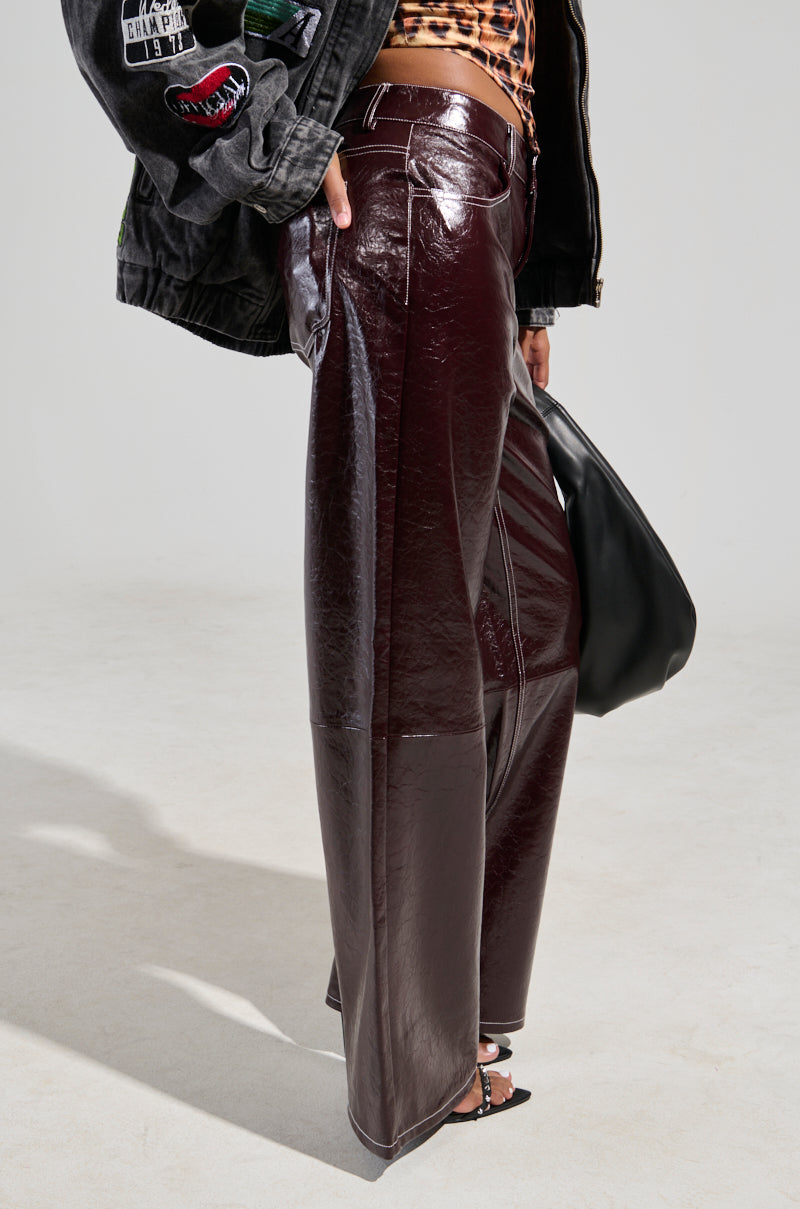 The individual is wearing the START WITH ME FAUX LEATHER TROUSER IN BURGUNDY, teamed with a black jacket adorned with patches and designs. They are also holding a black bag and sporting strappy sandals against a plain grey background. 2