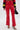 Someone dressed in SEXY FAUX LEATHER RED LACE-UP PANTS, a black, red, and yellow sports jersey, and black high-heeled sandals holds a small black handbag while standing against a plain white background.