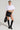 A person wearing a white puffy crop top and the TINA MINI SKIRT, which is a curve-hugging faux leather mini skirt, stands confidently. They hold a black bag and wear knee-high black boots paired with hoop earrings and bracelets, posing against a plain background.