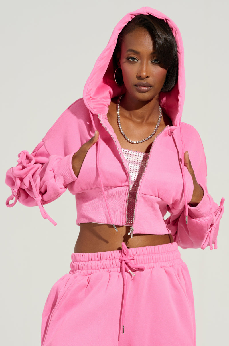 Dressed in the AKIRA Label TIE ME UP SWEATSHIRT, a person wears its pink cropped hoodie featuring drawstring details along with matching joggers, exuding confidence. With a hoop earring in one ear, the hood is up to showcase a sparkling top beneath this coquette ensemble against a plain background.
