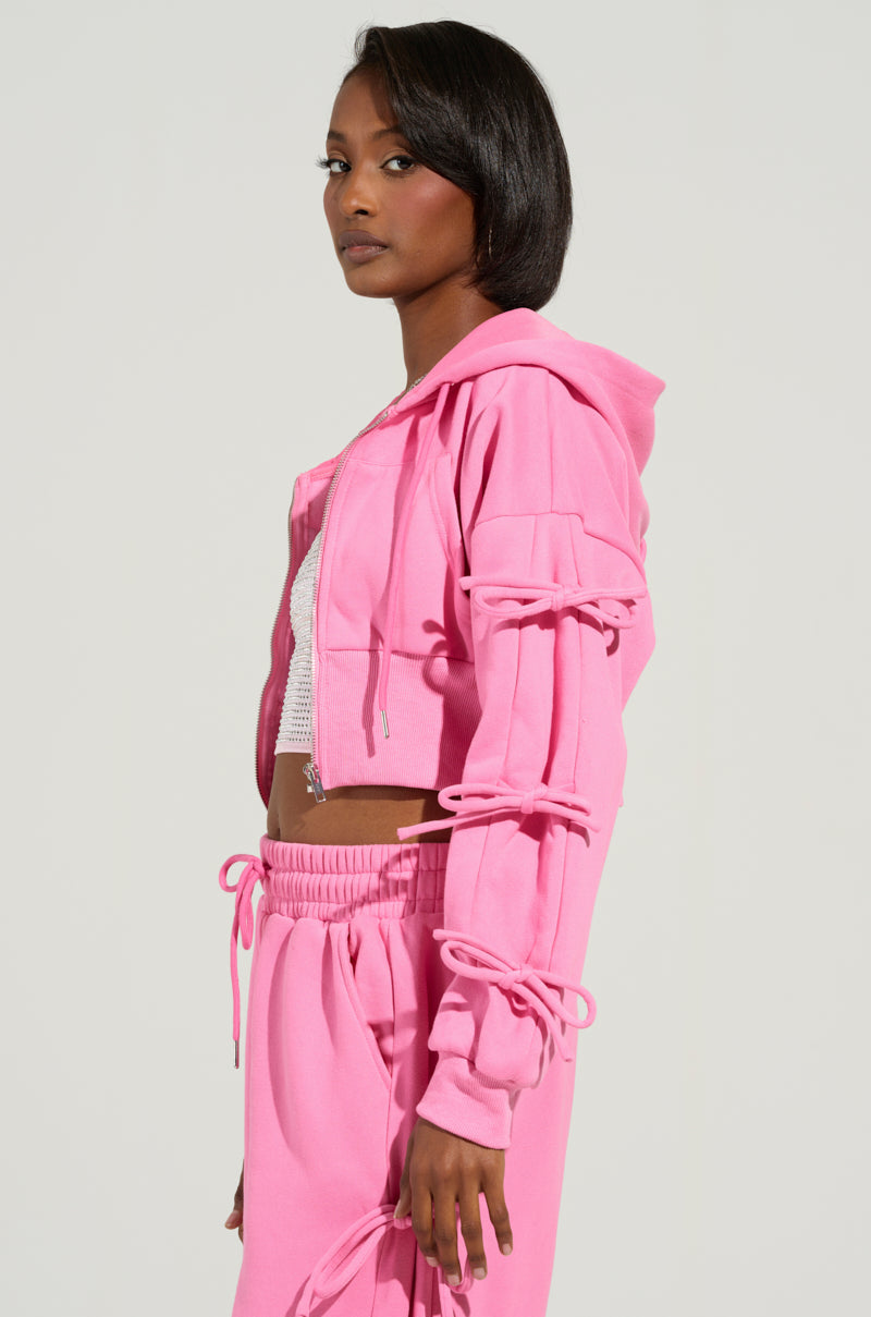 The person is wearing the TIE ME UP SWEATSHIRT from the AKIRA Label, featuring a coquette-style pink hoodie with lace-up sleeves, complemented by matching joggers. Their short hairstyle gives a contemporary touch as they gaze to the side against a simple background.