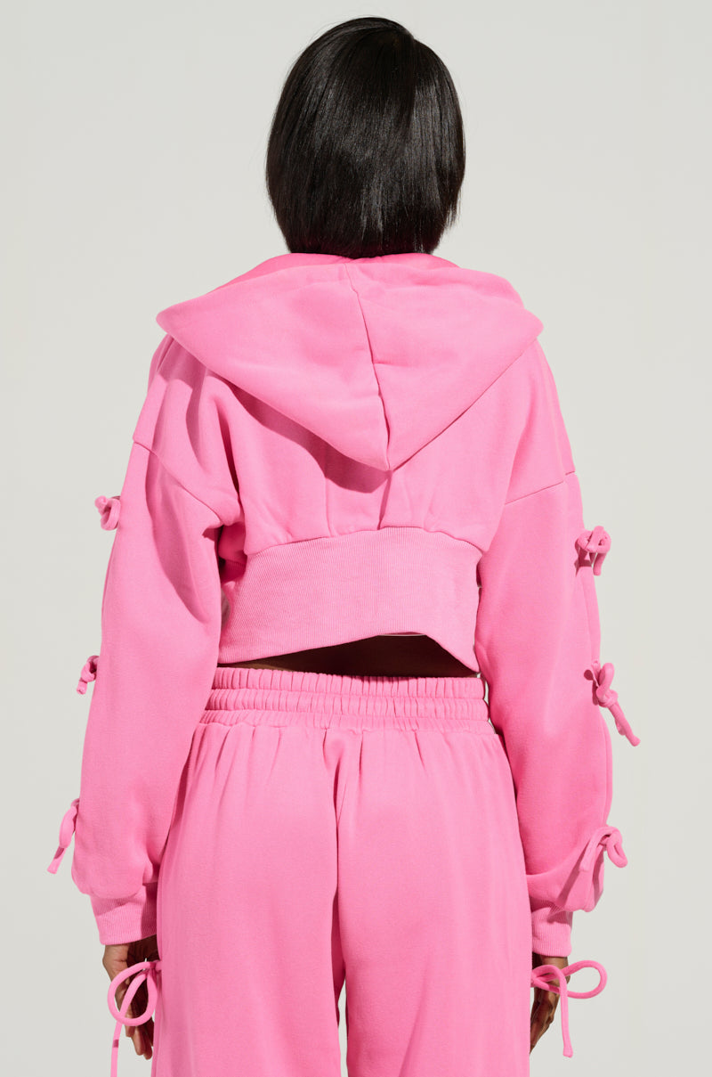 Dressed in the AKIRA Label's TIE ME UP SWEATSHIRT, this ensemble features a coquette charm with its pink hue and matching pants. The outfit is accented by bow details adorning the sleeves and sides, set against a soft backdrop as they turn away from the camera.
