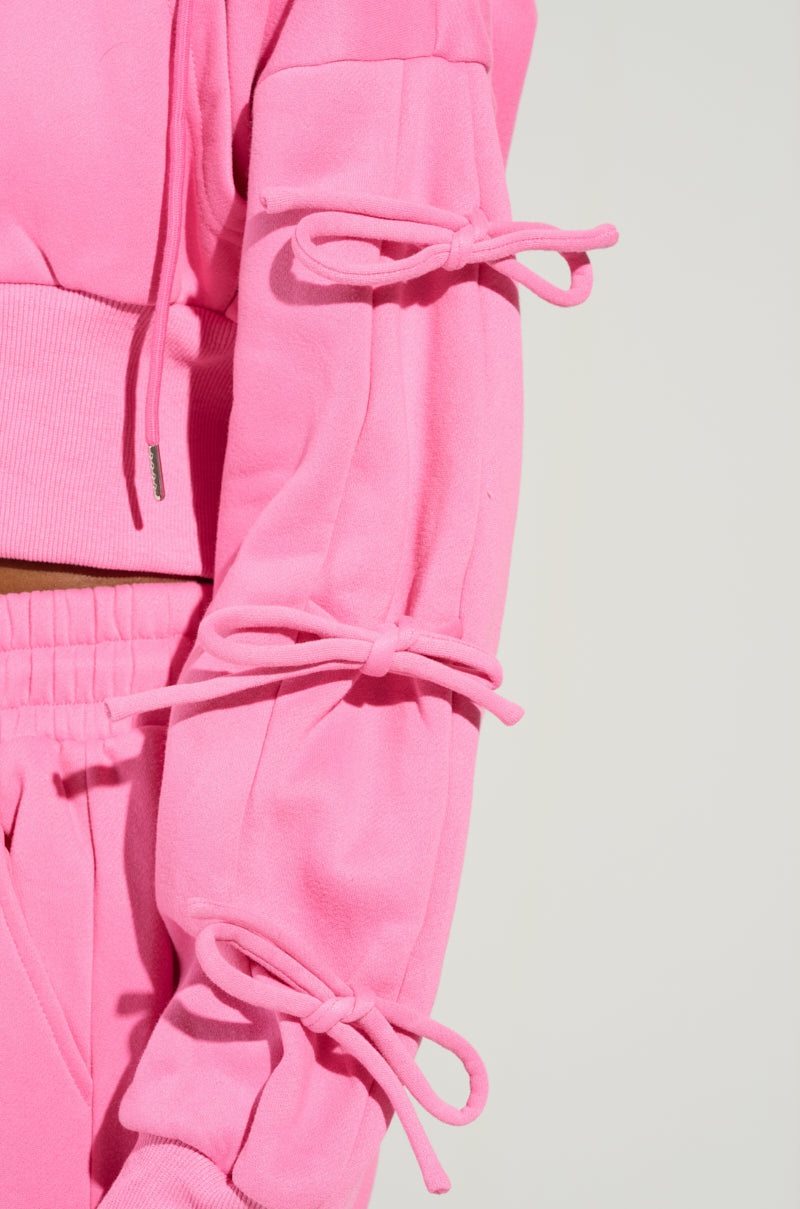 A close-up image highlights a stylish piece from the AKIRA Label, featuring the vibrant TIE ME UP SWEATSHIRT in bright pink. The attention is drawn to its long sleeves, embellished with three bows running along the arm. The fabric seems to be a cozy, substantial material, likely fleece or cotton.