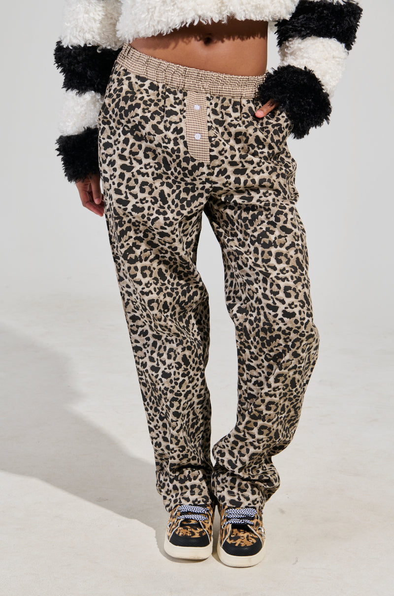 A person wearing the KISS ME CHEETAH PANT, featuring a stylish wide-leg silhouette, and a sweater with black-and-white fuzzy sleeves stands with one hand in their pocket. They pair the outfit with checkered pattern sneakers against a plain, light-colored background.