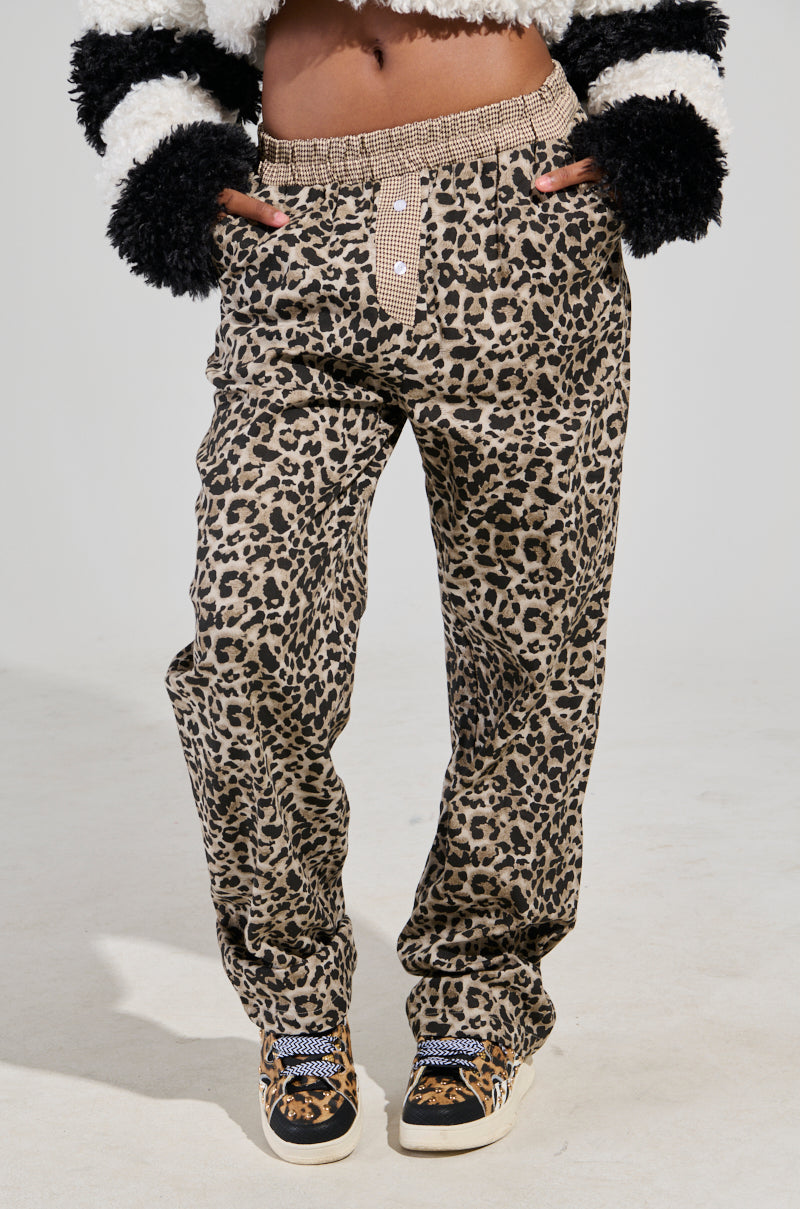 A person wearing KISS ME CHEETAH PANT and a fuzzy black and white sweater rests their hands in their pockets, complemented by patterned sneakers. The attention is on the clothing, with a plain background accentuating the wide leg silhouette of the pants.