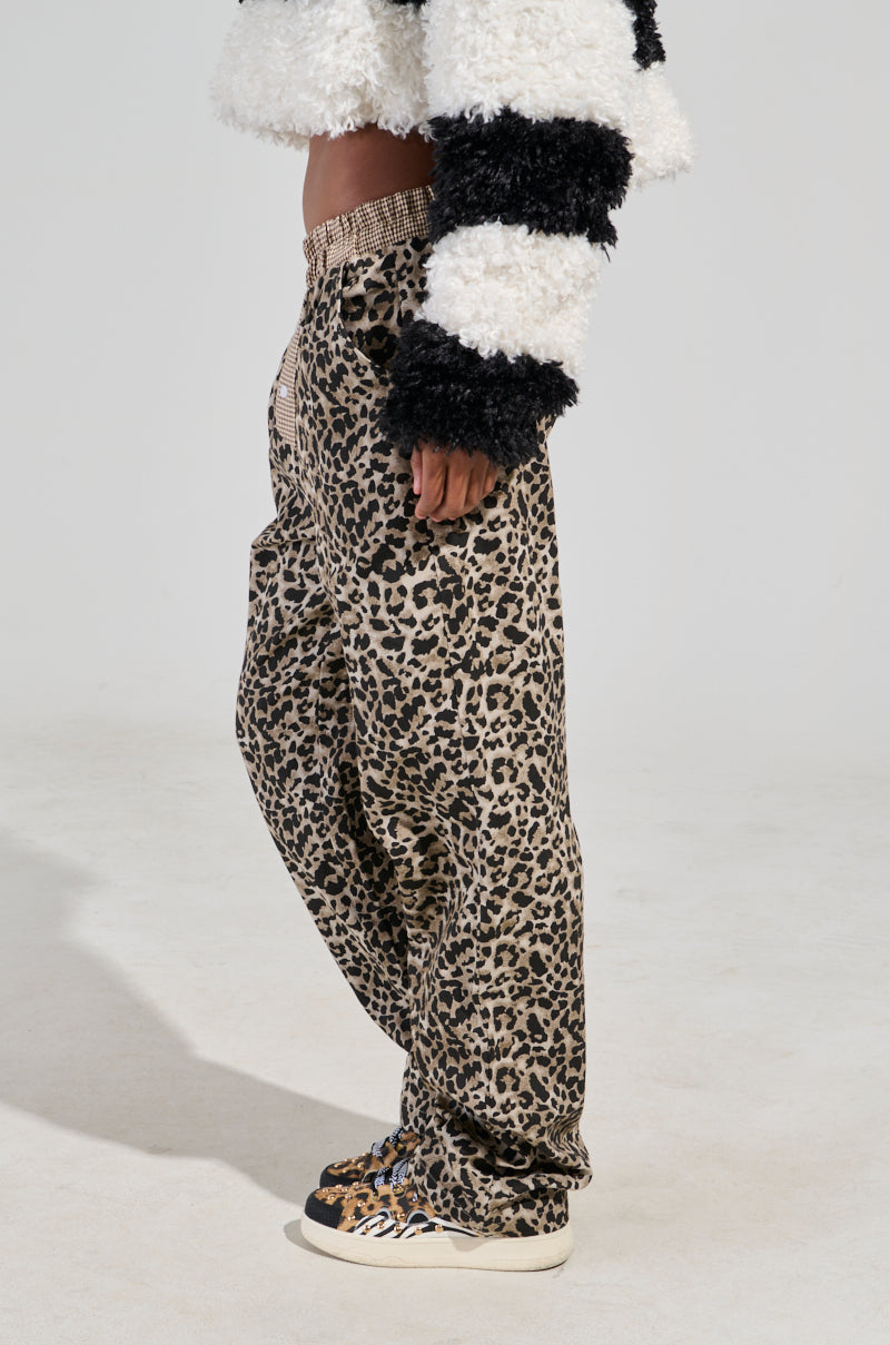 A person wearing the KISS ME CHEETAH PANT with a wide leg silhouette and matching sneakers stands sideways. They are also wearing a fluffy black and white striped sweater against a plain, light-colored background.