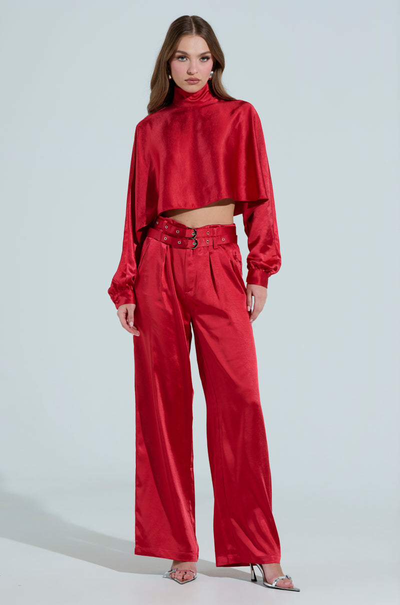 A person showcases the FEELIN THE LOVE BELTED SATIN TROUSER—high-rise with a double belt—paired with a red, long-sleeve cropped top. They have long hair and open-toed heels, standing against a neutral background.