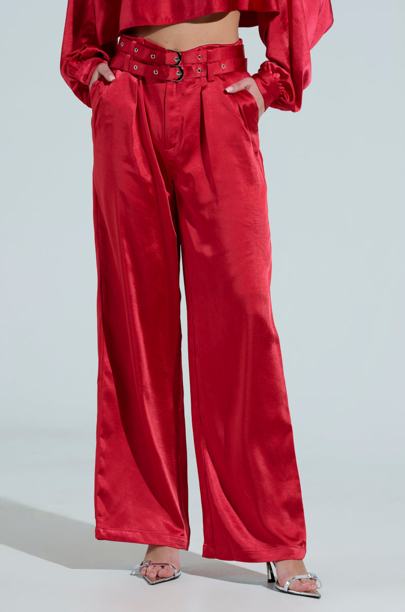 Someone shows off FEELIN THE LOVE BELTED SATIN TROUSER in red with a matching cropped top, hands in pockets. The wide-leg satin pants are teamed with strappy silver high heels against a simple light backdrop.