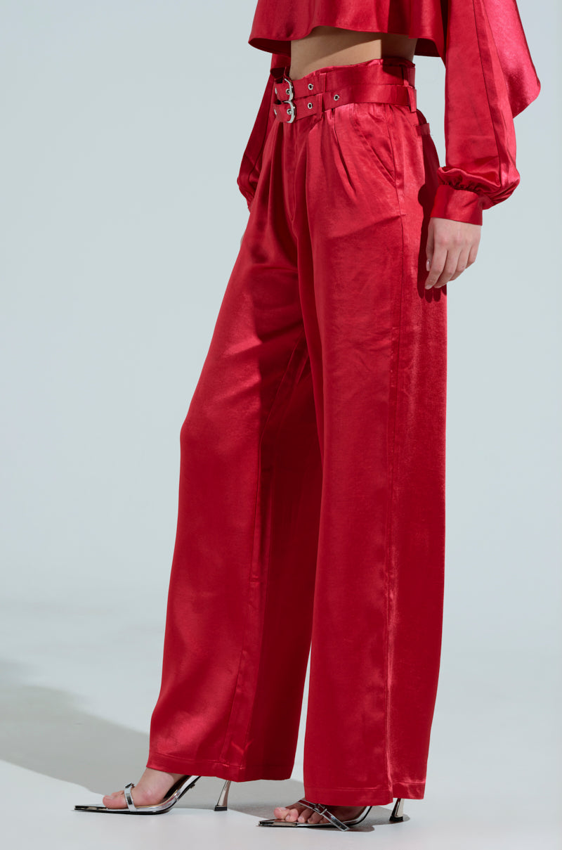 A person in a red satin outfit wears a cropped long-sleeve top with FEELIN THE LOVE BELTED SATIN TROUSER, featuring a double belt design, paired with pointed, translucent high-heeled shoes against a light gray background.