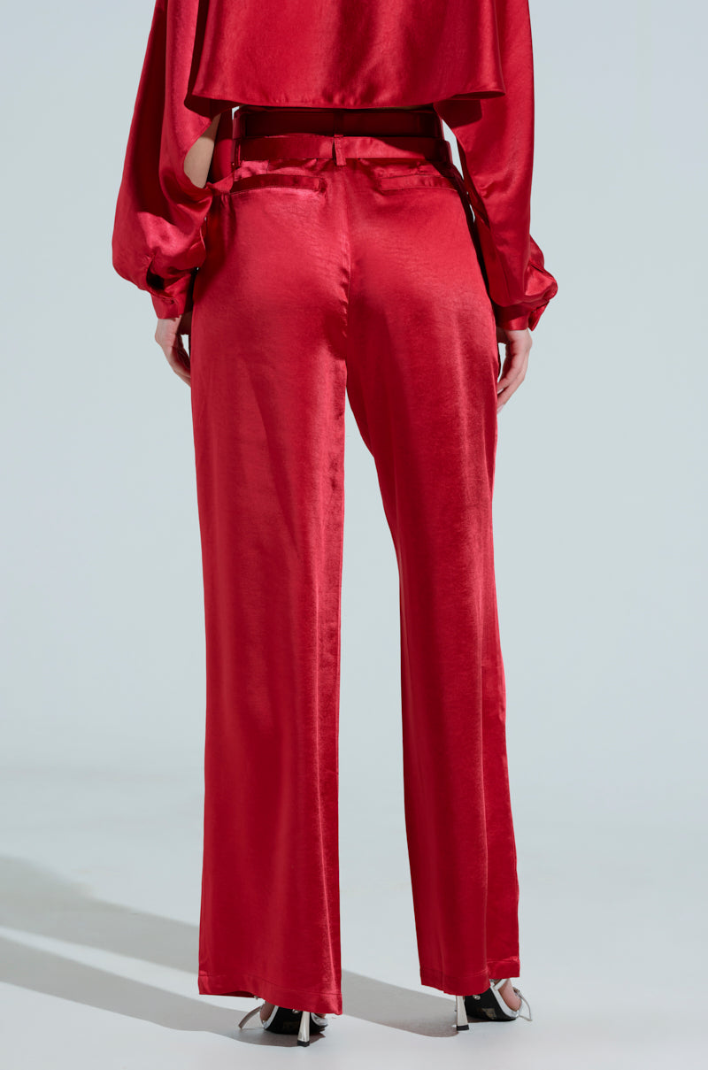 Rear view of a person in a shiny red outfit, featuring the FEELIN THE LOVE BELTED SATIN TROUSER with a high-rise waist and a matching top. The sleek ensemble is paired with black and white shoes against a plain, light background.