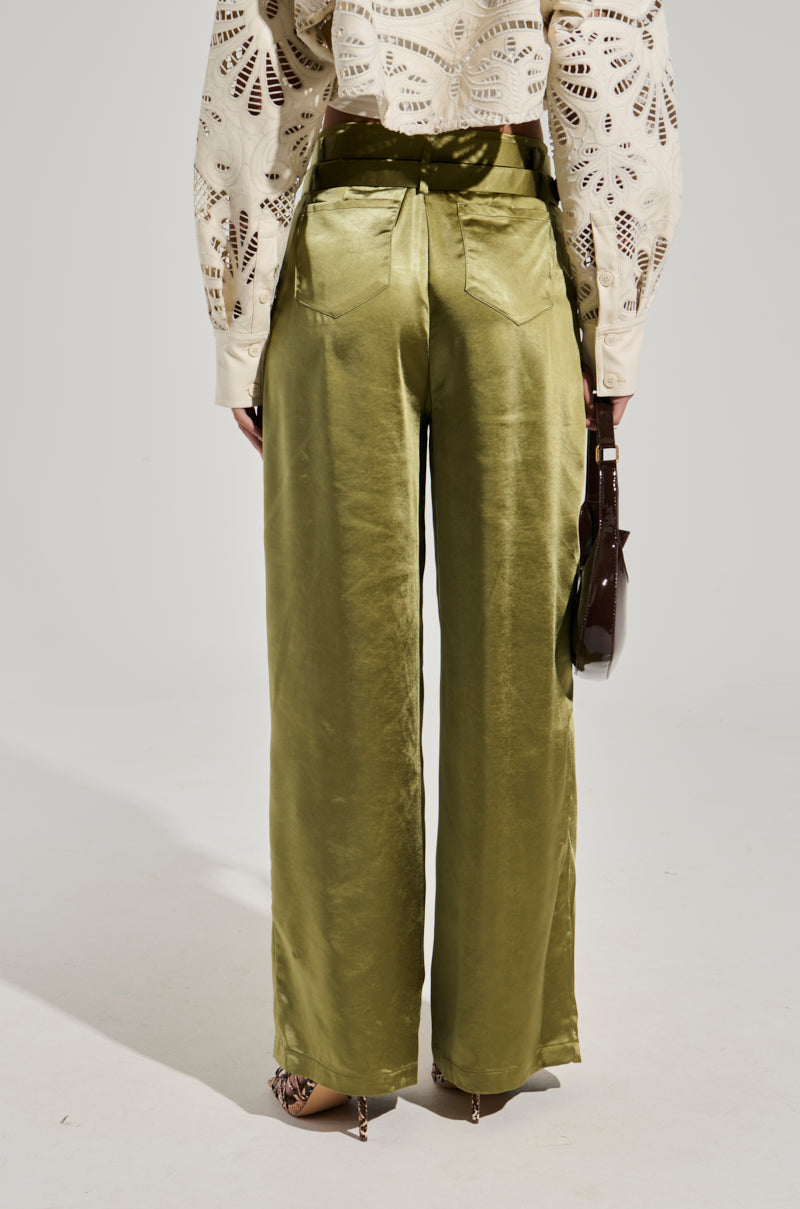 A person stands facing away, wearing the SLICK AND SWEET TROUSER in olive green satin paired with a cream-colored, long-sleeved top featuring a cutout pattern. They hold a small brown handbag by their side, and their shoes are partially visible. The background is plain and light-colored.