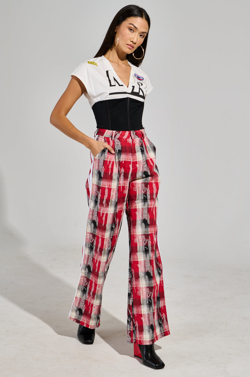 A woman model poses in the CRAZY LIKE ME PLAID TROUSER, featuring a chic wide-leg silhouette and high-rise cut, with her hands tucked into the pockets. She's styled in a white T-shirt layered under a black corset and accessorized with hoop earrings. Her neutral expression complements her ensemble, which is finished off with sleek black ankle-length boots.