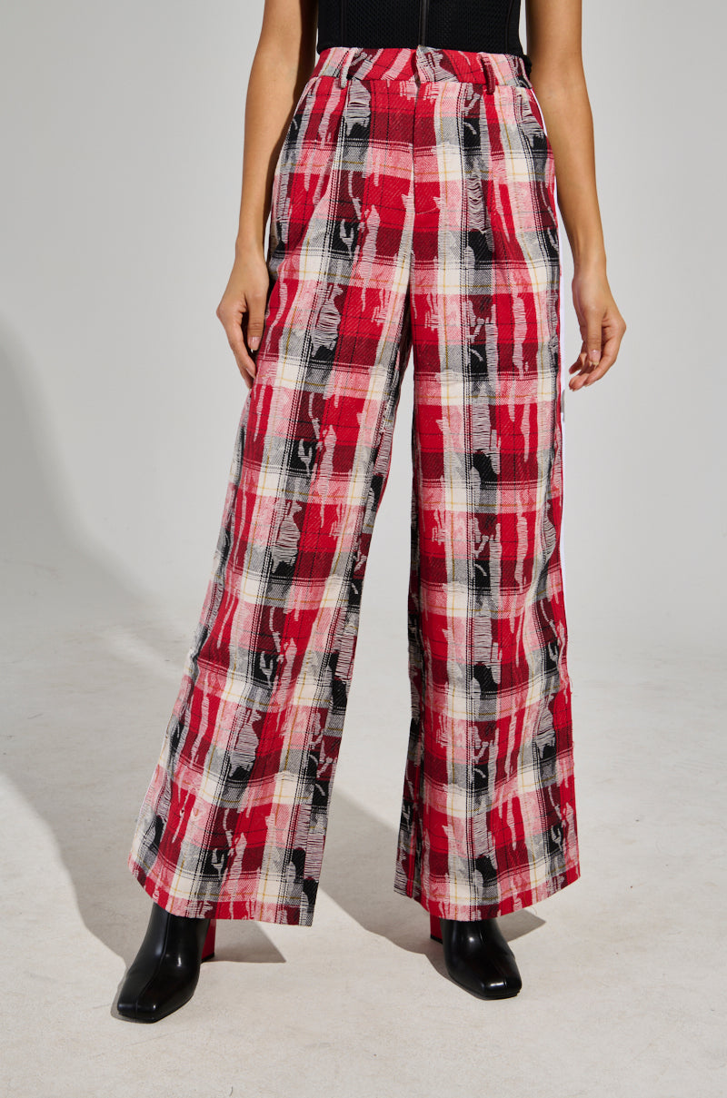 A person models the CRAZY LIKE ME PLAID TROUSER, showcasing its high-rise design with a red, black, and white plaid pattern. The trousers pair flawlessly with sleek black boots against a plain gray background, while their upper body remains intriguingly out of view.