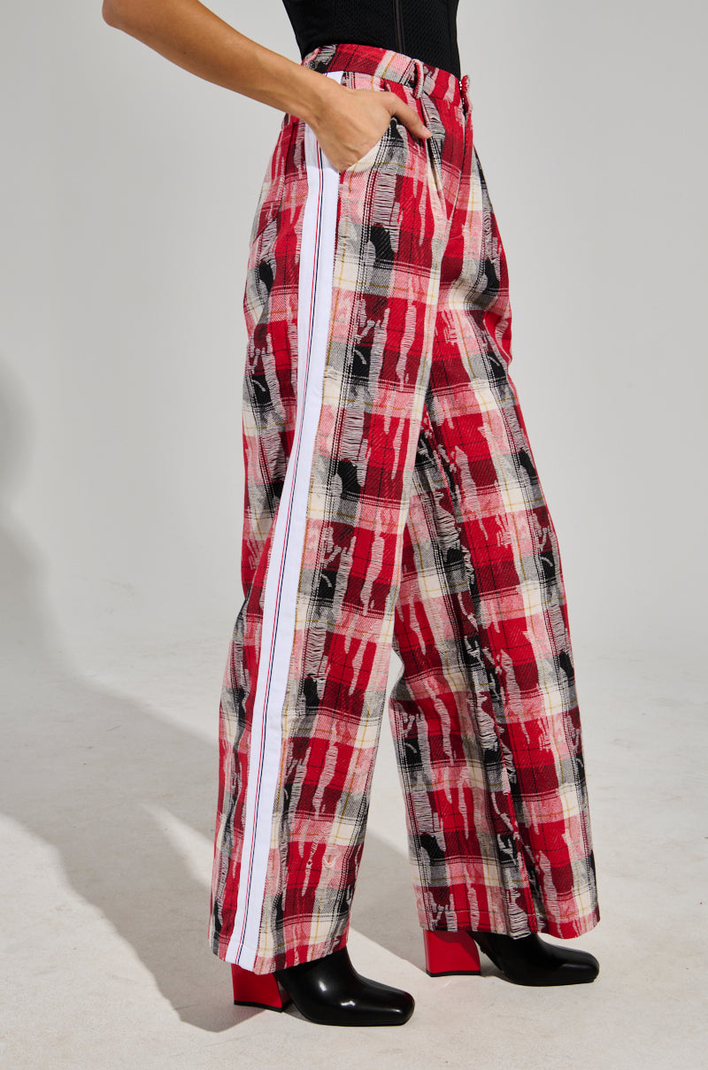 A person wearing the CRAZY LIKE ME PLAID TROUSER featuring a red and black plaid pattern with white side stripes and black boots stands on a light background. The high-rise design offers a loose fit, with one hand casually tucked in a pocket.