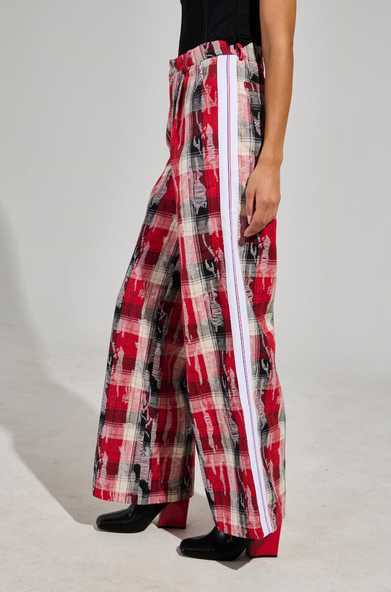 The person is sporting the chic "CRAZY LIKE ME PLAID TROUSER" in a red plaid pattern with a white stripe down the side, complemented by black shoes. The wide-leg silhouette pops against the simple light gray backdrop.