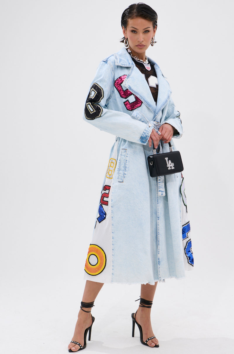 A woman exudes sporty-chic flair in the COUNTDOWN PATCH DENIM TRENCH, a light blue denim coat detailed with varsity-style patches and numbers, paired with black strappy heels and a small black LA handbag. 0