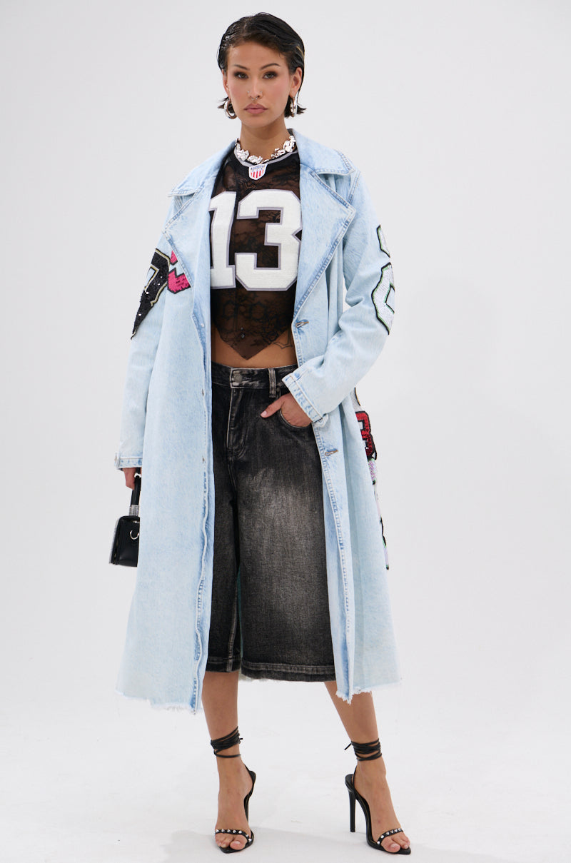 A woman models the COUNTDOWN PATCH DENIM TRENCH over a black mesh football jersey with “13,” wide-leg black denim shorts, and black heels, creating a sporty-chic vibe while holding a small black purse against a white background. 1