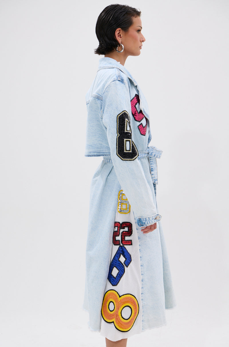A woman with short dark hair models the COUNTDOWN PATCH DENIM TRENCH, a light blue coat featuring large varsity-style patches. She stands sideways, facing right, against a plain white background. 2