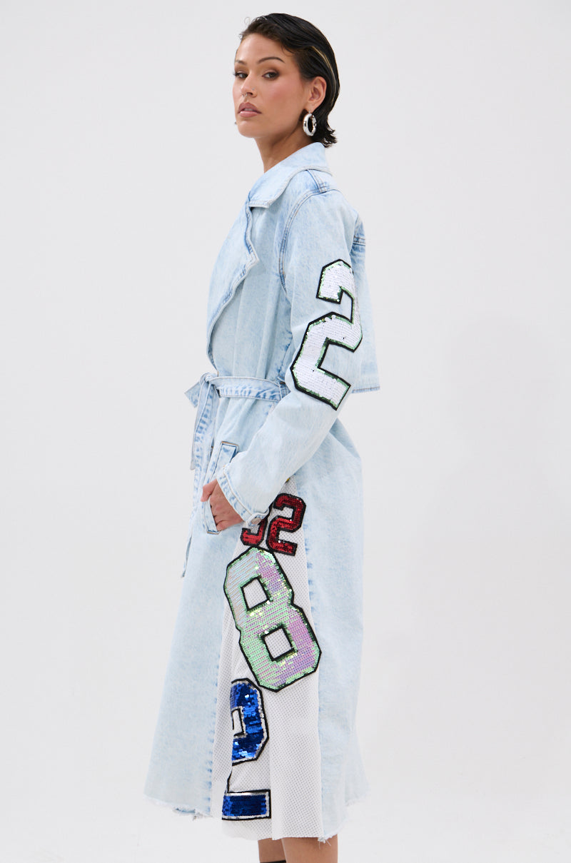 A woman with short hair and large hoop earrings poses sideways against a white background in the COUNTDOWN PATCH DENIM TRENCH, featuring varsity-style colorful sequin patches on the sleeves and side for a sporty-chic look. 3